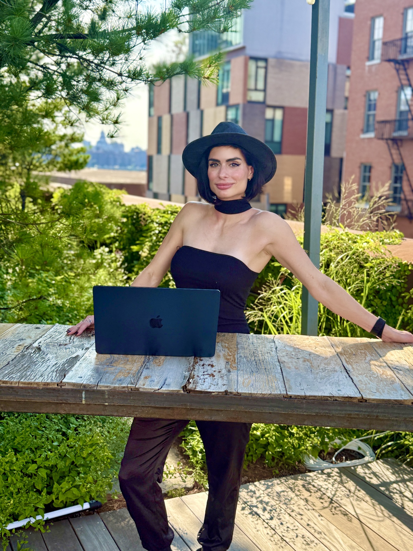Carina in a black hat at a table in a New York garden, her laptop open in front of her